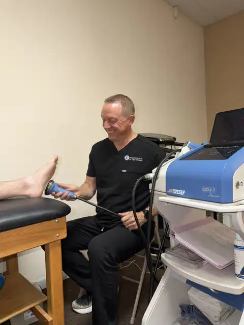 Healthcare provider performing shockwave therapy on a patient’s foot and ankle using a handheld device while the patient sits on a treatment table in a chiropractic clinic.